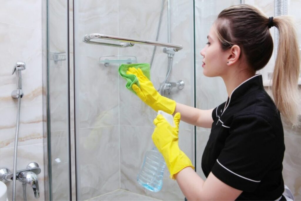 a woman wearing gloves spraying vinegar on shower door then wiping it with cloth