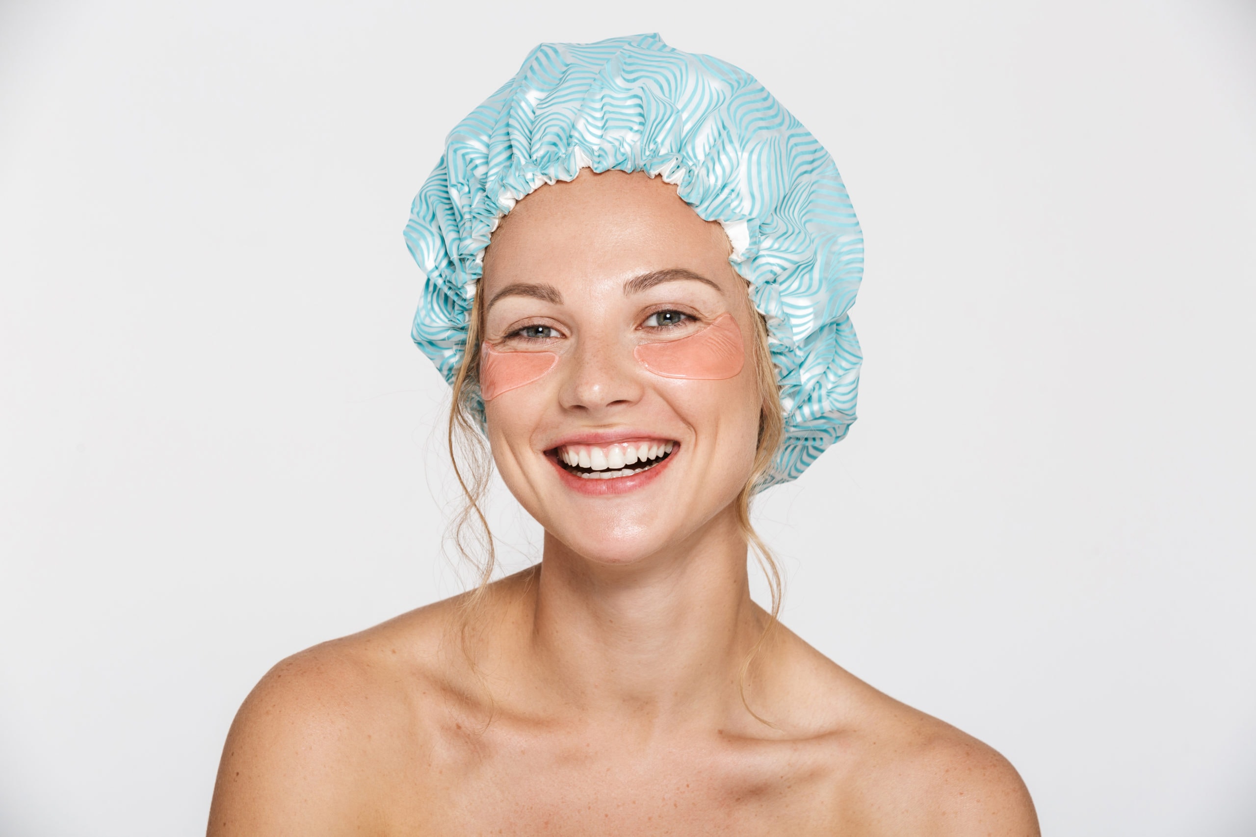 Image of happy half-naked woman wearing cosmetic patches and shower cap laughing isolated over white background