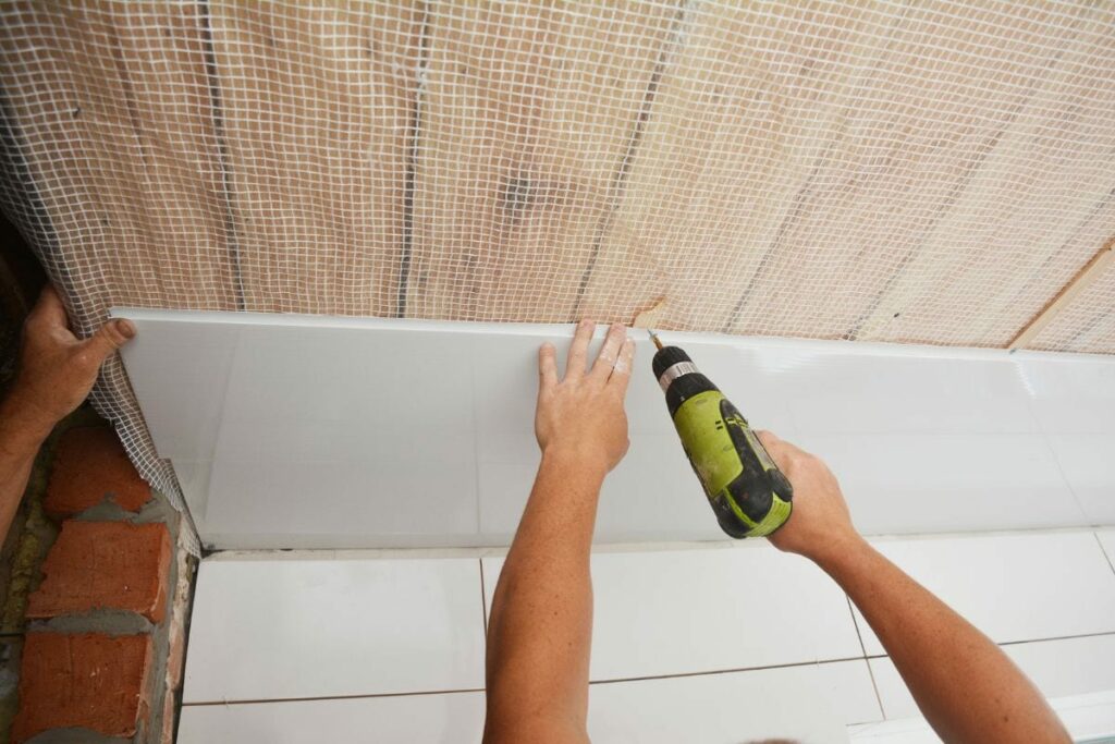 A builder is installing plastic ceiling panel, white PVC bathroom ceiling cladding in a bathroom using an electric screwdriver to attach it to the planked wood ceiling with a vapor barrier.