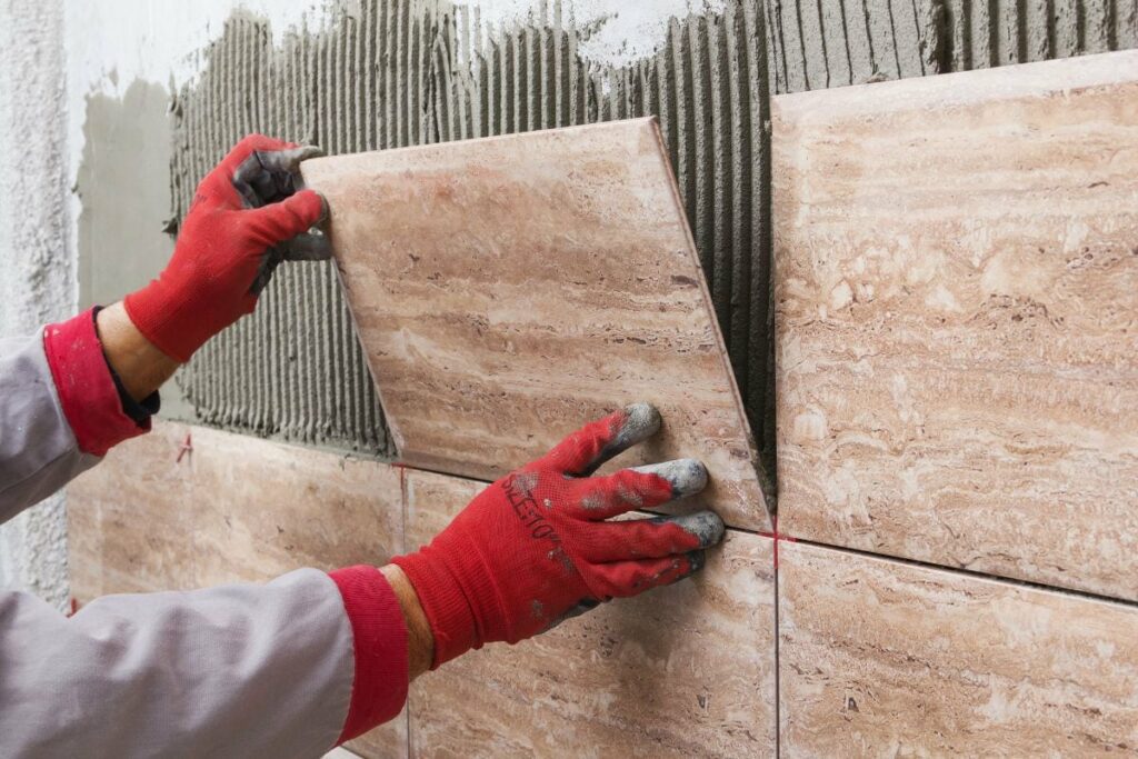 Close-up of worker tiler hands installing light beige ceramic tiles on walls of future bathroom